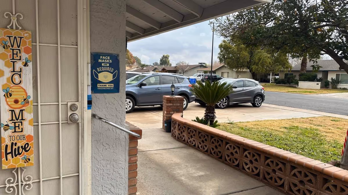 View from the entrance of Beloved Care Home, LLC showing a welcome sign with bees and honeycomb design on the door, a sign indicating face masks are required, a small brick wall with decorative blocks, a palm plant, and a driveway with parked cars. Residential houses and trees are visible in the background under a cloudy sky.