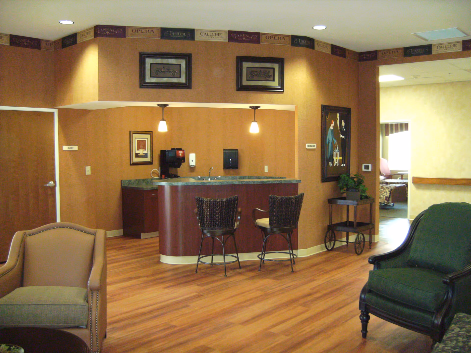 Interior view of a senior living facility lounge area featuring a small kitchenette with two pendant lights, a countertop with two wicker bar stools, framed artwork on the walls, and comfortable armchairs on a wood floor. There is a doorway leading to another room with a bed visible in the background.