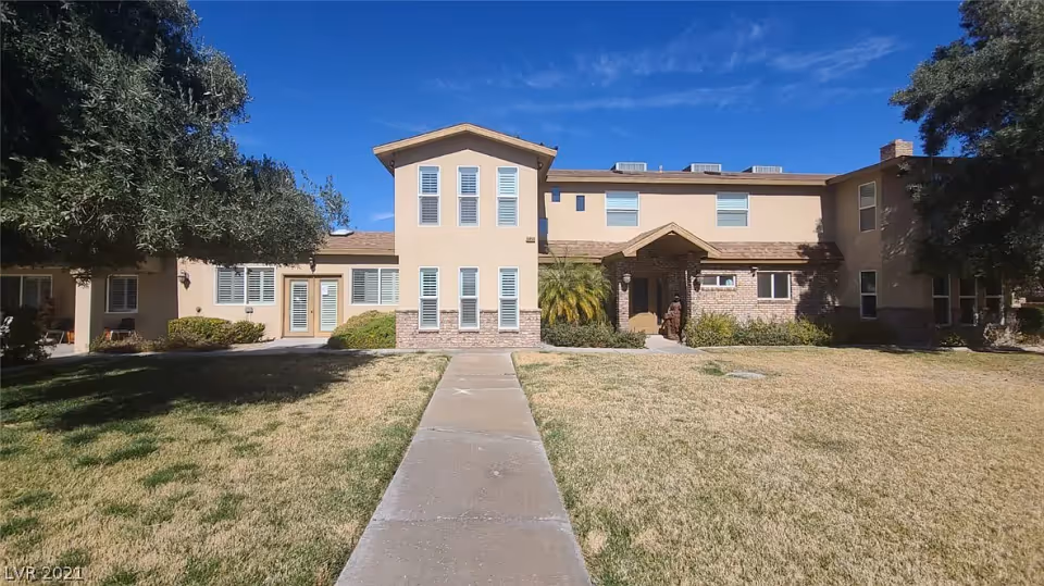 Front exterior view of a two-story beige residential building with multiple windows and a central entrance with a small porch. There is a concrete walkway leading up to the entrance, surrounded by a lawn with some trees and bushes. The sky is clear and blue.