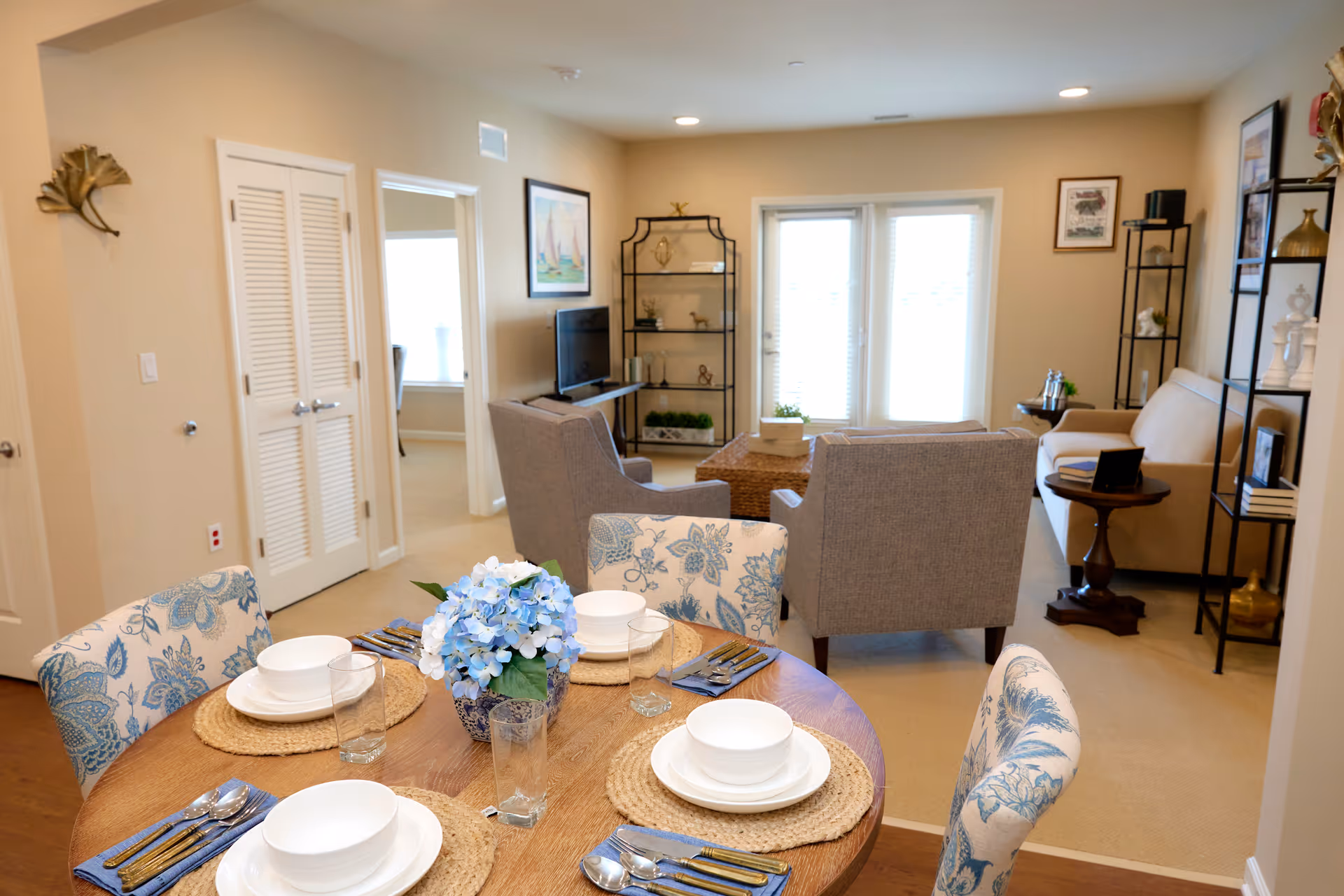 A cozy living and dining area in a senior living facility. The foreground shows a round wooden dining table set with white dishes, glasses, silverware, and a blue floral centerpiece. The dining chairs have a blue and white floral pattern. In the background, there are two gray armchairs facing a TV on a black metal shelf, a beige sofa, and additional shelving units with decorative items. French doors with blinds allow natural light into the room. The walls are painted beige and decorated with framed artwork.