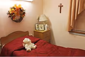 Single-occupancy bedroom with a red bedspread, a stuffed animal on the bed, a nightstand holding a lamp and framed photos, and wall decorations including a cross and a wreath.