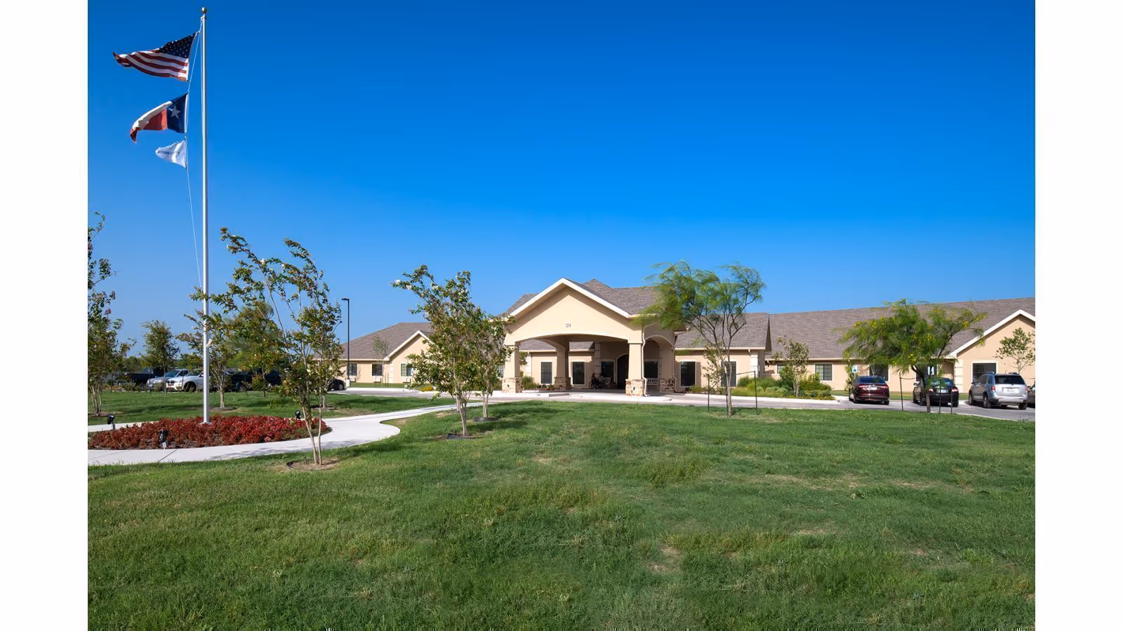 Front exterior of a single-story beige senior living facility with a circular driveway, flagpoles, parked cars, and a large grassy lawn under a clear blue sky.