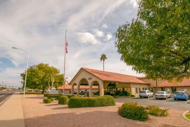 Exterior view of Citadel Post Acute facility with a tiled roof entrance, an American flag on a flagpole, several parked cars, and landscaped bushes and trees under a partly cloudy sky.