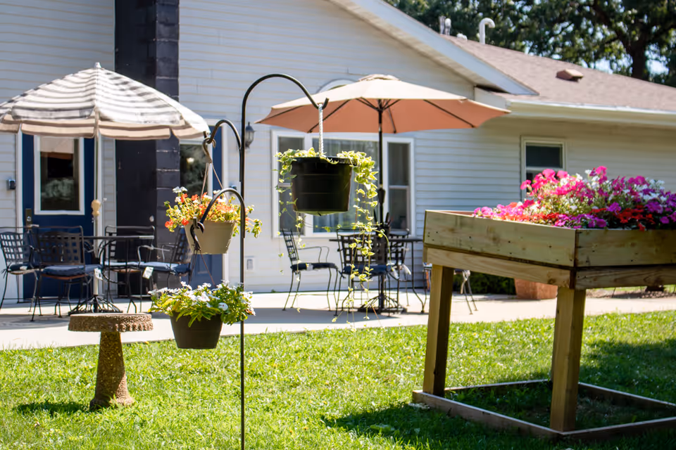Outdoor patio area with green grass, hanging flower pots, a raised wooden flower bed filled with colorful flowers, and metal tables and chairs under umbrellas in front of a white building.
