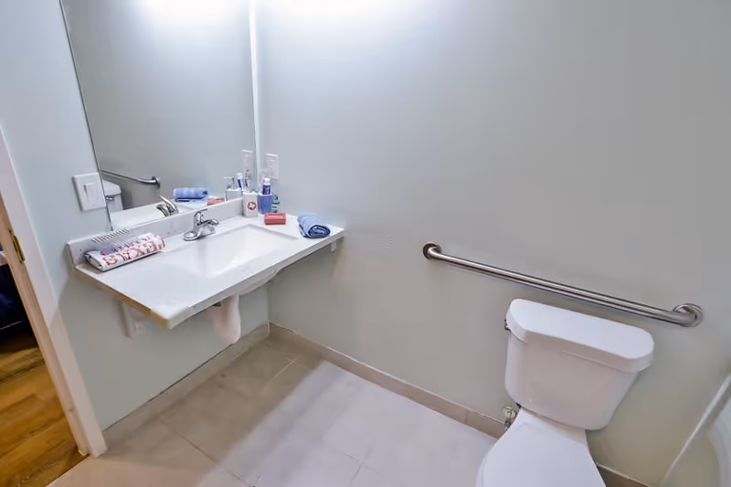 A clean and accessible bathroom with a white toilet, a wall-mounted sink with a faucet, a large mirror above the sink, and a grab bar installed on the wall next to the toilet. Towels and toiletries are neatly placed on the sink counter.