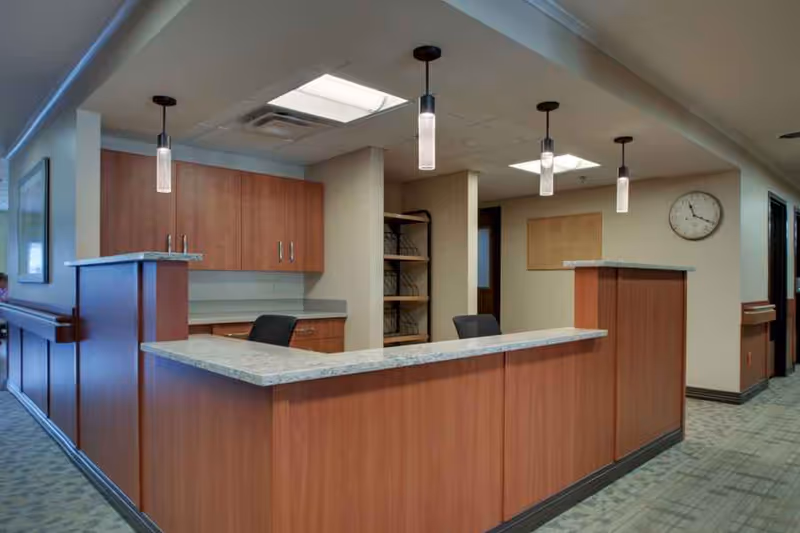 Reception desk area inside a senior living facility with wood paneling and granite countertops, three pendant lights hanging from the ceiling, office chairs behind the desk, a wall clock, and a bulletin board on the wall.