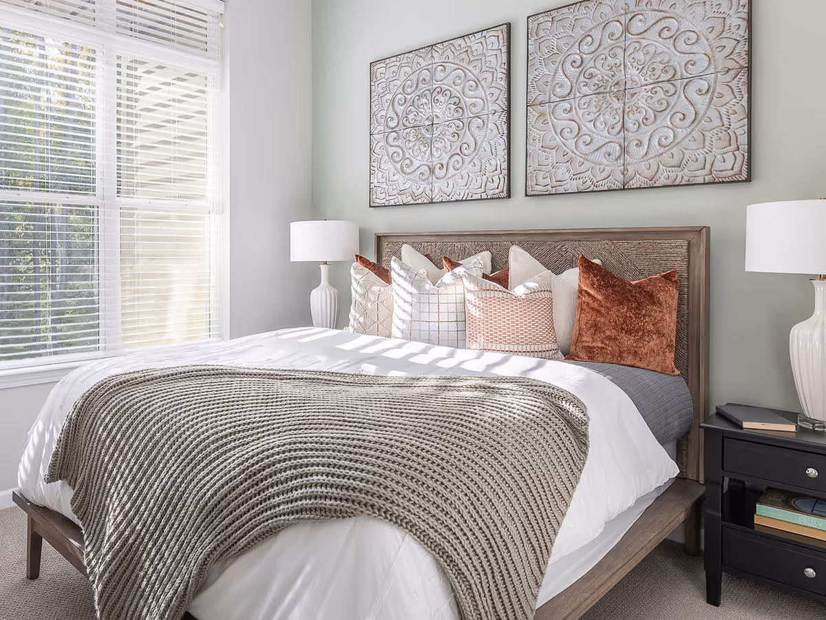A bright bedroom with a large bed featuring multiple decorative pillows and a knitted throw blanket. Two white table lamps sit on black nightstands on either side of the bed. Above the bed are two large, intricately carved wall art pieces. A window with white blinds allows natural light to fill the room.