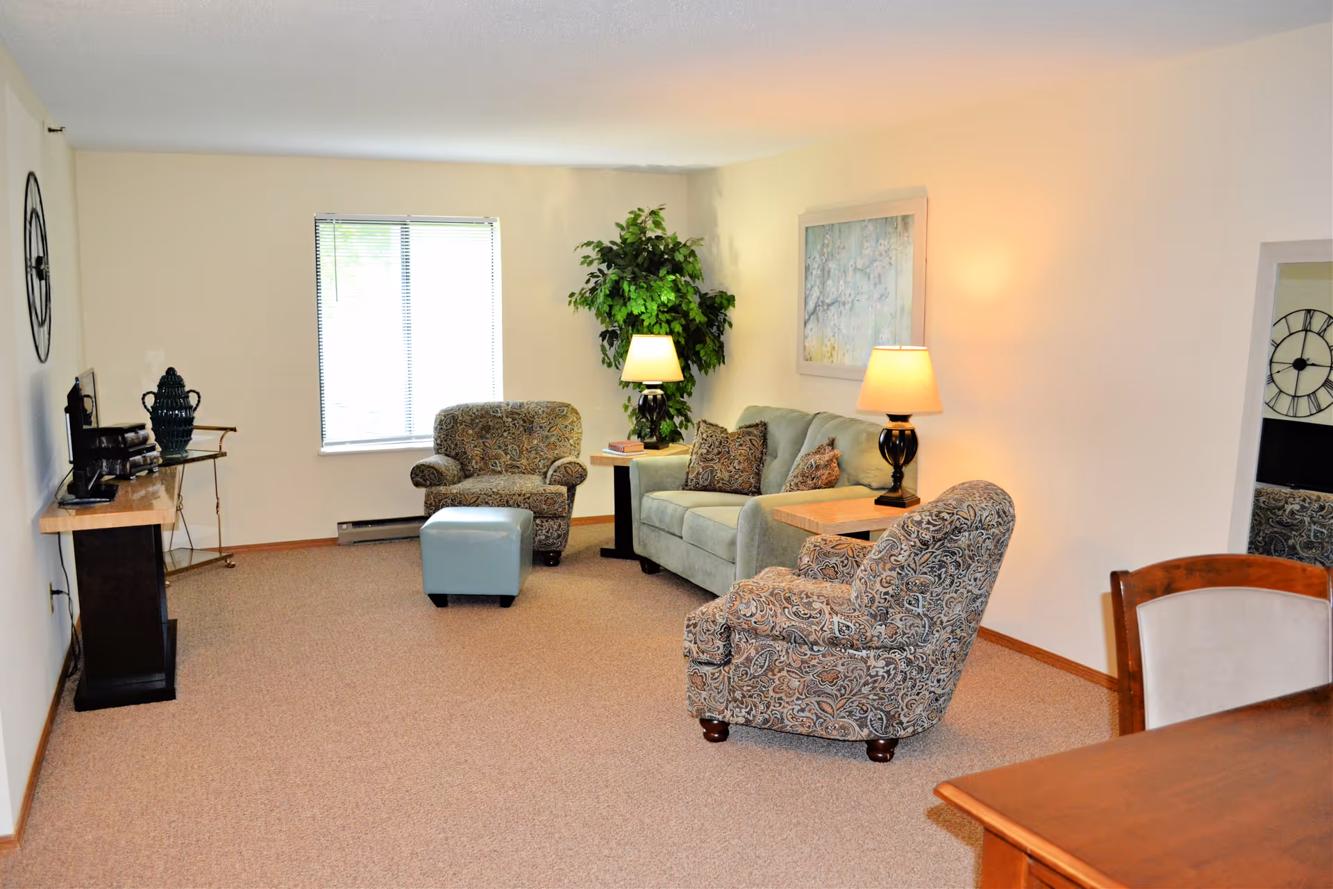 Bright living room with a sofa, two patterned armchairs, ottoman, lamps, TV stand, and wall art.
