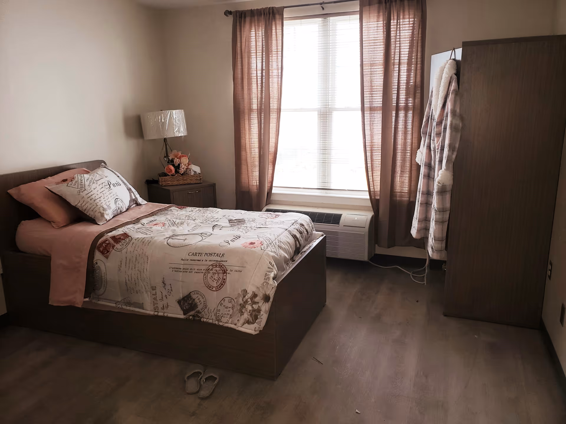 A cozy bedroom with a single bed covered in a Paris-themed bedspread and pink sheets. Next to the bed is a nightstand with a lamp and a basket of flowers. A window with sheer brown curtains lets in natural light. A wooden wardrobe stands to the right with two robes hanging on its side. A pair of slippers is placed on the wooden floor near the bed.