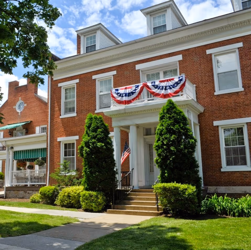 Exterior view of a red brick building with white trim and columns at the entrance. The building has a small balcony decorated with red, white, and blue bunting. There are two tall evergreen bushes flanking the entrance steps, and an American flag is displayed near the door. The sky is partly cloudy and there is a green lawn and sidewalk in front.