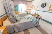 A nursing center room with two hospital-style beds, each made with blankets and pillows. The room has a window with green curtains, a clock on the wall, framed pictures, and a small table with flowers and decorative items between the beds.