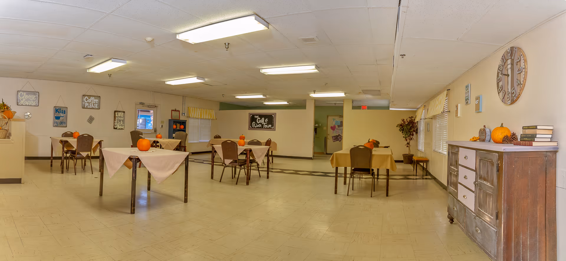 A spacious dining room in a senior living facility with several tables covered with beige tablecloths, each decorated with a small pumpkin. The room has beige walls adorned with framed signs and decorations, a large clock on the right wall, and a wooden sideboard with books and seasonal decor. Fluorescent ceiling lights illuminate the room.