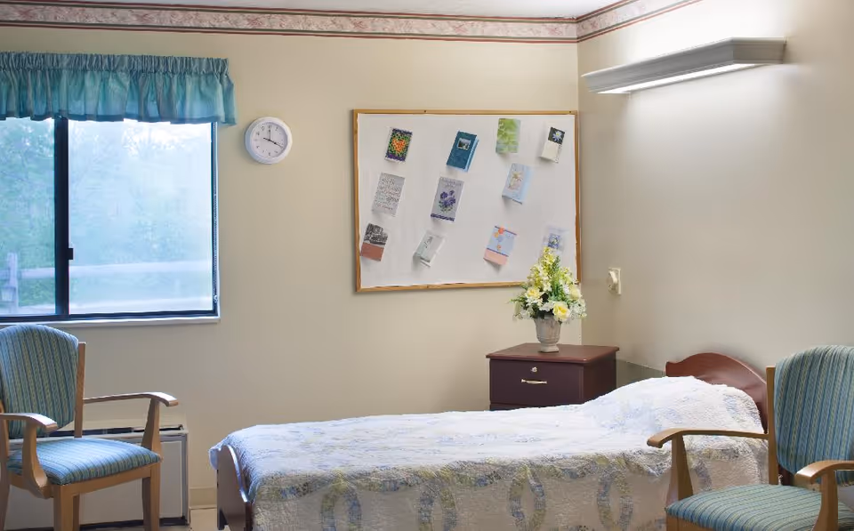 A simple bedroom in a senior living facility with a single bed covered with a white quilt, two wooden chairs with blue striped cushions, a nightstand with a flower vase, a bulletin board with various papers pinned on the wall, a clock, and a window with a blue valance.