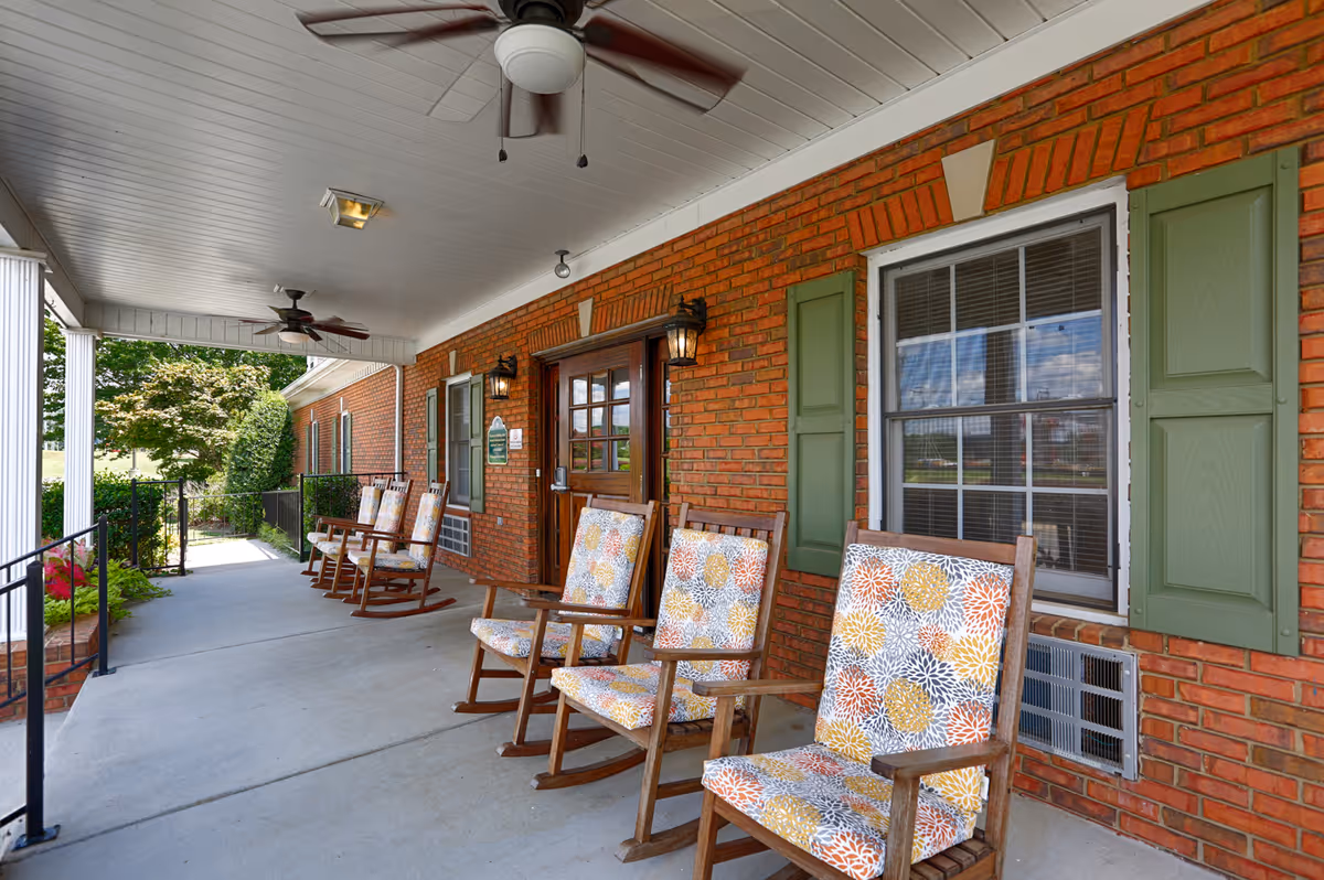 Covered outdoor porch area with a row of wooden rocking chairs featuring colorful floral cushions. The porch has a white ceiling with ceiling fans and lights, red brick walls with green shutters on the windows, and a wooden door. There are plants and trees visible beyond the porch railing.