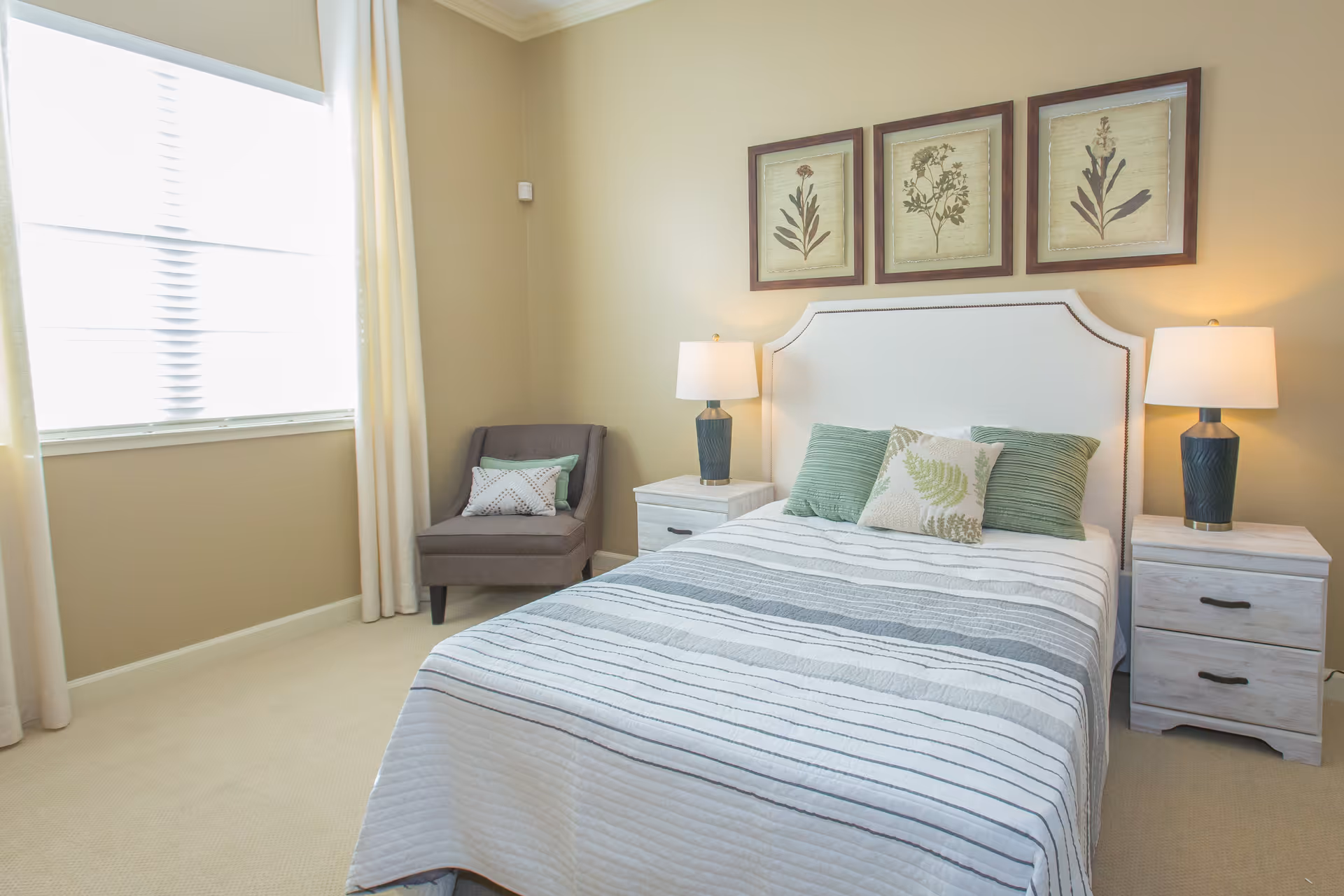 A bright bedroom with a bed featuring a white headboard and striped bedding in shades of gray and white. There are three decorative pillows on the bed, two green and one with a leaf pattern. On either side of the bed are white nightstands with drawers, each topped with a blue lamp with a white shade. A gray armchair with two pillows is positioned near a large window with white blinds and cream-colored curtains. Above the bed are three framed botanical prints.