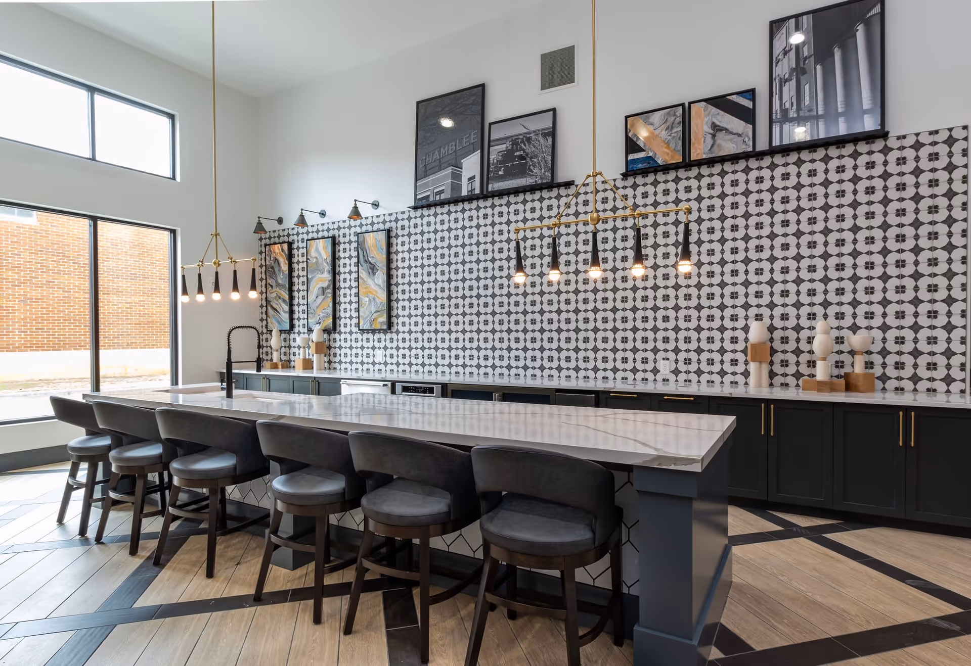 Modern kitchen area with a long marble island countertop surrounded by six dark cushioned bar stools. The back wall features black and white patterned tiles, black lower cabinets with gold handles, and decorative artwork. Two gold pendant light fixtures hang above the island. Large windows allow natural light to fill the space.