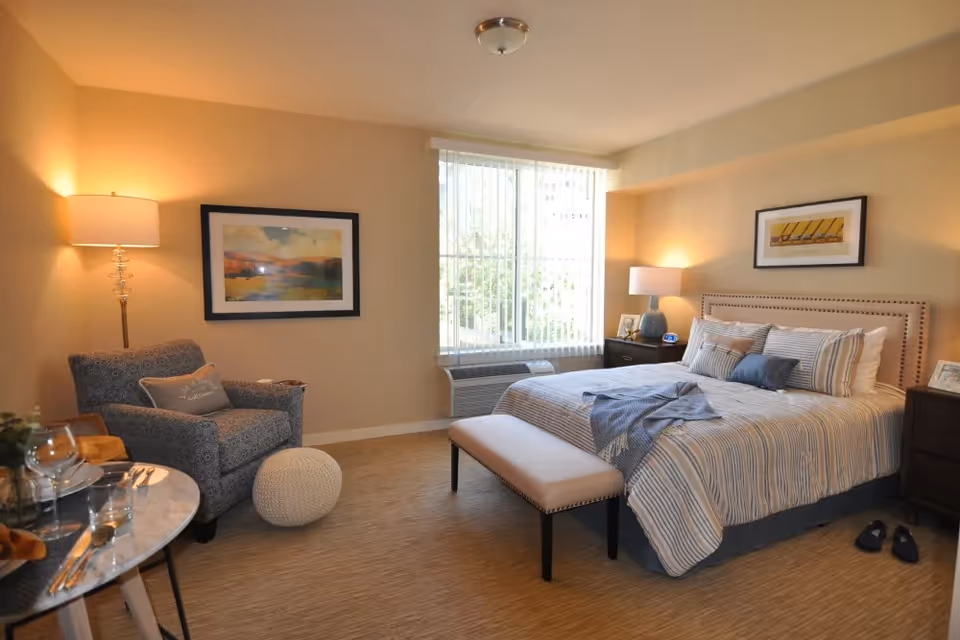 A cozy bedroom with a large bed dressed in striped bedding and multiple pillows. There is a cushioned bench at the foot of the bed, two dark wooden nightstands with lamps, and framed artwork on the walls. A comfortable armchair with a pillow and a round knitted ottoman are placed near a small round table set with glassware and napkins. A large window with vertical blinds lets in natural light.