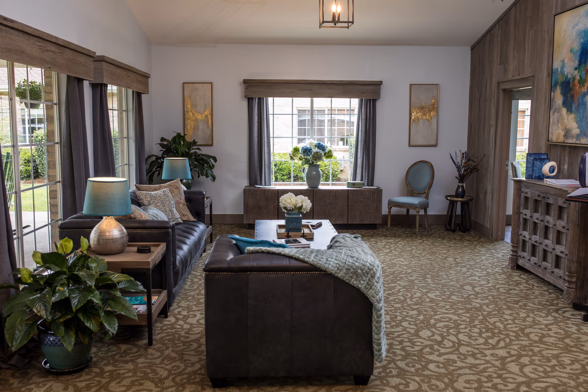 A cozy living room with two dark leather sofas, a wooden coffee table with books and flowers, a side table with a lamp, potted plants, a large window with curtains, a wooden cabinet, an upholstered chair, and decorative artwork on the walls.
