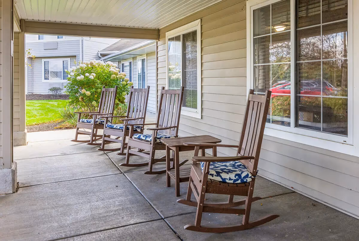 Covered outdoor patio area with four wooden rocking chairs with blue and white patterned cushions, a small wooden table, beige siding walls, and windows reflecting a red car and greenery outside.