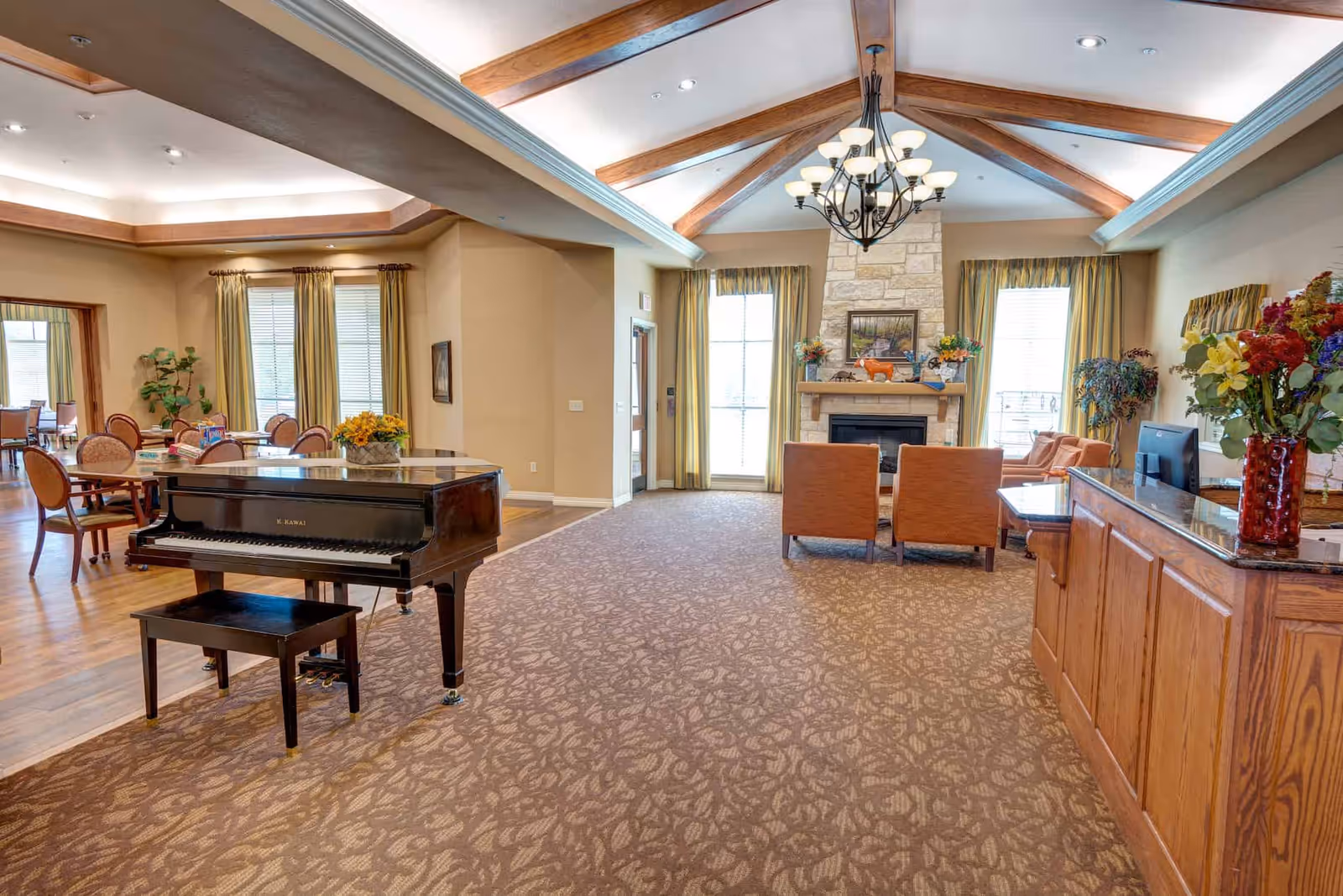 A spacious senior living facility common area with a grand piano on the left, a seating area with two orange chairs facing a stone fireplace in the center, and a wooden reception desk with a vase of flowers on the right. The room features high vaulted ceilings with wooden beams, large windows with curtains, and a carpeted floor with a patterned design.
