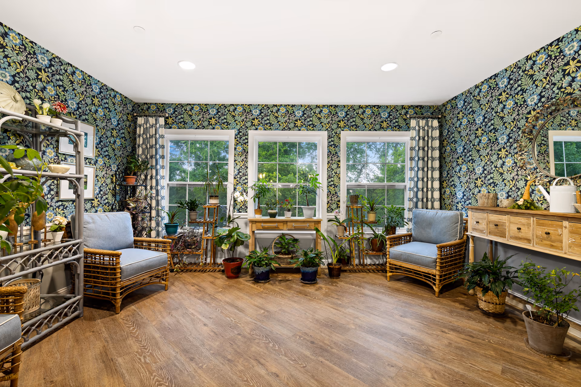 A bright and cozy sitting room with floral patterned wallpaper and three large windows letting in natural light. The room features two wicker armchairs with blue cushions, several potted plants on the floor and on shelves, a wooden console table with drawers, and a round mirror above it. The floor is wooden, and the overall atmosphere is inviting and homey.