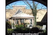 View through an archway of an outdoor courtyard area with a gazebo, surrounded by greenery and outdoor seating, under a clear sky.
