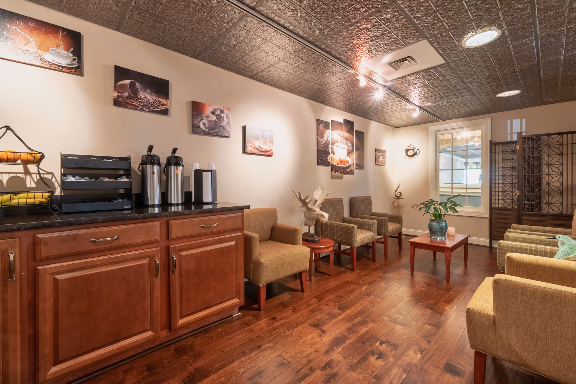 A cozy seating area with multiple armchairs arranged around a small wooden coffee table with a plant on it. The room has wooden flooring and a decorative ceiling with recessed lighting. On the left side, there is a countertop with coffee dispensers, cups, and a rack with supplies. The walls are decorated with coffee-themed artwork and a window with a view into another room.