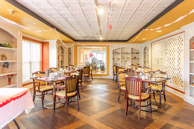 A bright dining room with several wooden tables and chairs arranged neatly. The tables are set with cups, glasses, and silverware. The room features large windows with blinds, built-in shelves with decorative items, and a patterned wooden floor. Streamers and star-shaped decorations hang from the ceiling.
