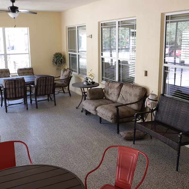 A covered outdoor patio area with multiple seating options including cushioned chairs, a loveseat, a bench, and a dining table with chairs. The floor is tiled, and there are large windows along the wall. A ceiling fan is mounted above, and a small round table with decorative items is placed near the loveseat.