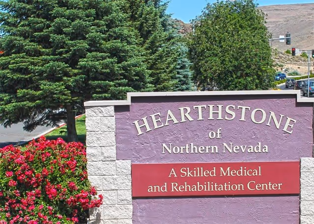 Outdoor sign for Hearthstone of Northern Nevada, a skilled medical and rehabilitation center, surrounded by green trees and red flowers with a clear blue sky in the background.