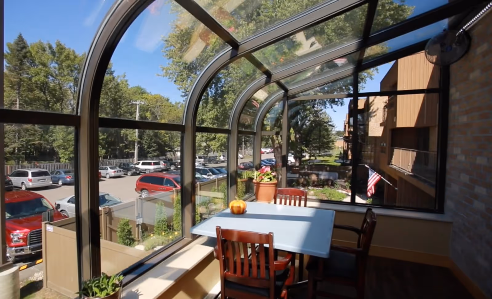 A sunroom with large curved glass windows overlooking a parking lot and trees. Inside the sunroom is a table with four wooden chairs, a small pumpkin decoration on the table, and a potted plant. The exterior of a building and an American flag are visible outside the windows.