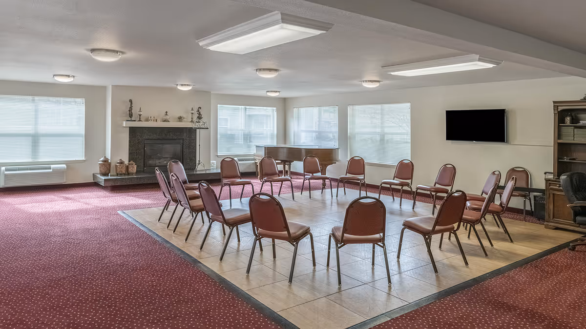 A senior living facility common room with a circle of empty chairs arranged on a tiled floor area surrounded by red carpet. The room features large windows with blinds, a fireplace with decorative items on the mantel, a grand piano, a wall-mounted TV, and a wooden cabinet with shelves.