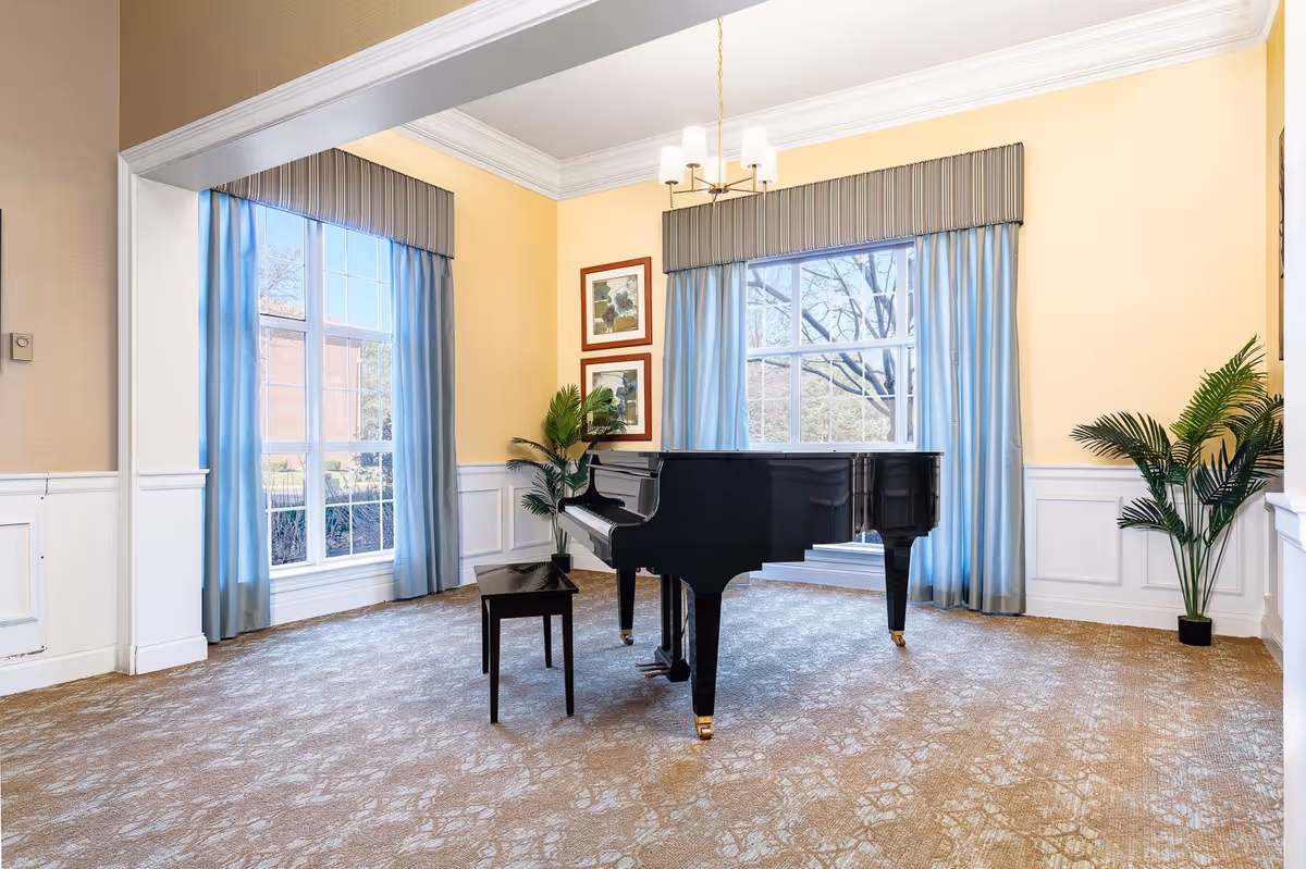 A bright room with yellow walls and large windows covered with blue curtains. In the center of the room is a black grand piano with a matching bench. The room has beige patterned carpet, white wainscoting, two framed pictures on the wall, a chandelier hanging from the ceiling, and two potted plants in the corners.