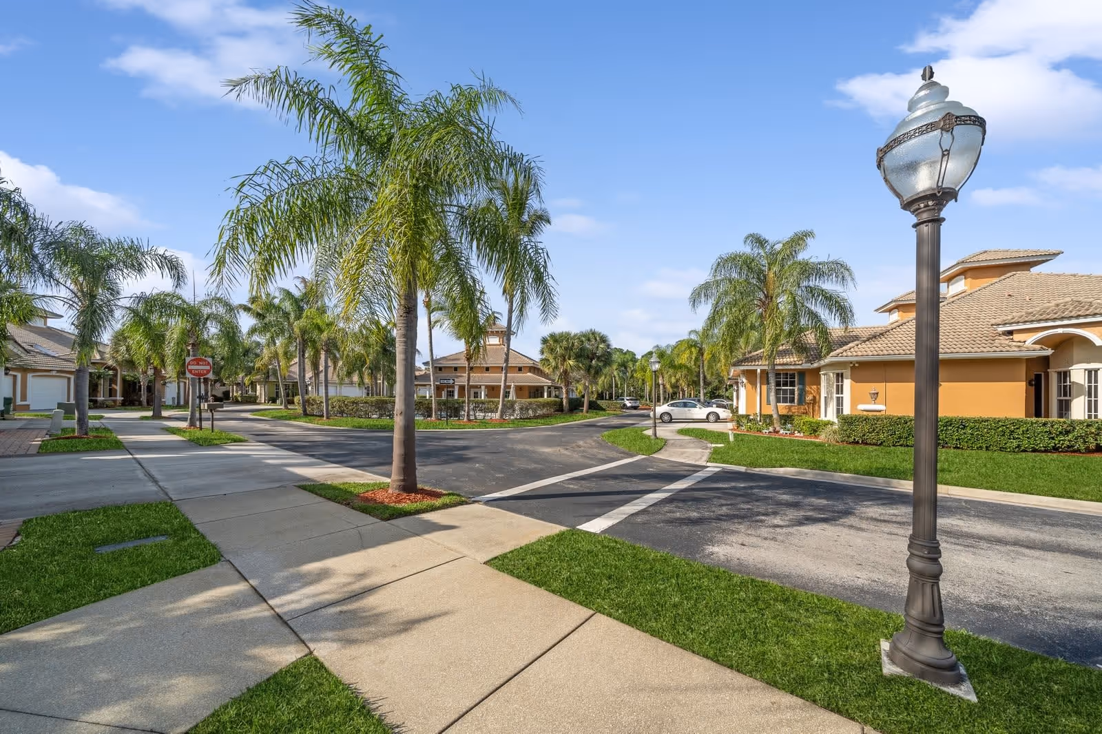 A sunny outdoor view of a residential community with palm trees lining the sidewalks and streets. Beige buildings with tiled roofs are visible, along with a vintage-style street lamp in the foreground. The sky is clear with a few clouds.