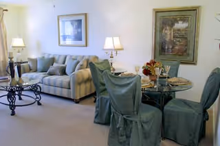 A cozy living and dining area featuring a striped sofa with multiple cushions, a glass coffee table with a decorative base, and a round glass dining table with four chairs covered in green fabric slipcovers. The room is softly lit by two table lamps and decorated with framed artwork on the walls.