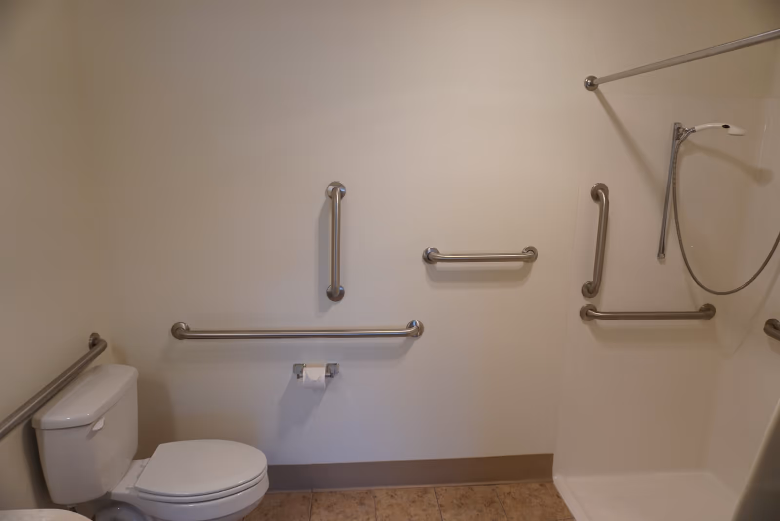 A bathroom with a toilet and a walk-in shower equipped with multiple stainless steel grab bars for accessibility. The walls are light-colored, and the floor has beige tiles.