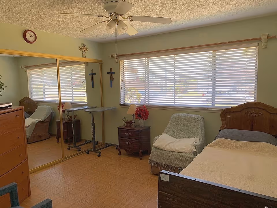 A bedroom with a single bed covered with a beige blanket, a cushioned armchair with a white pillow and green throw, a wooden nightstand with a lamp and a vase of red flowers, a mirrored closet door, a ceiling fan with lights, and a large window with horizontal blinds letting in natural light. The walls are light green and decorated with three crosses and a clock.