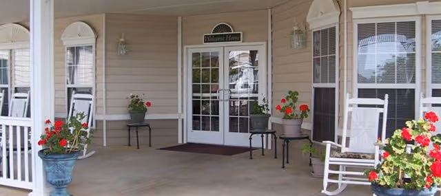 Covered entrance area of a building with double glass doors and a 'Welcome Home' sign above them. The porch has white rocking chairs, potted plants with red flowers, and beige siding on the walls.