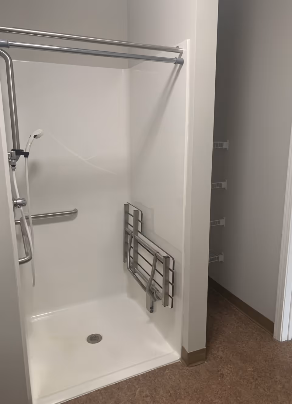 A white accessible shower stall with a handheld showerhead, grab bars, and a fold-down shower seat mounted on the wall. Adjacent to the shower is an open doorway leading to a small closet with wire shelving. The floor is a brownish color and the walls are painted light gray.