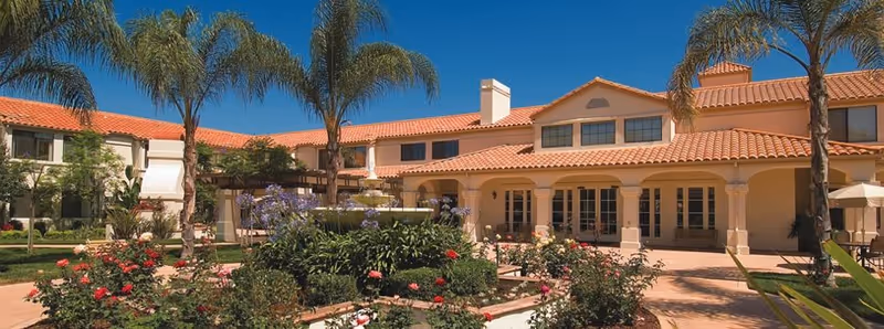 Exterior view of Oakmont of Chino Hills facility featuring a two-story building with a red tile roof, surrounded by palm trees and a garden with blooming flowers under a clear blue sky.