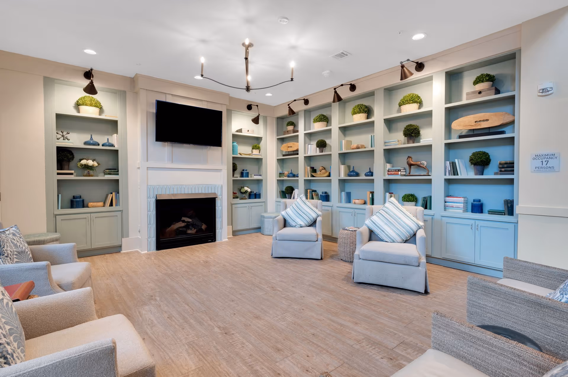 A cozy living room with light blue built-in shelves filled with decorative items and plants, a fireplace with a mounted flat-screen TV above it, and several cushioned armchairs with striped pillows arranged around the room on a wooden floor.
