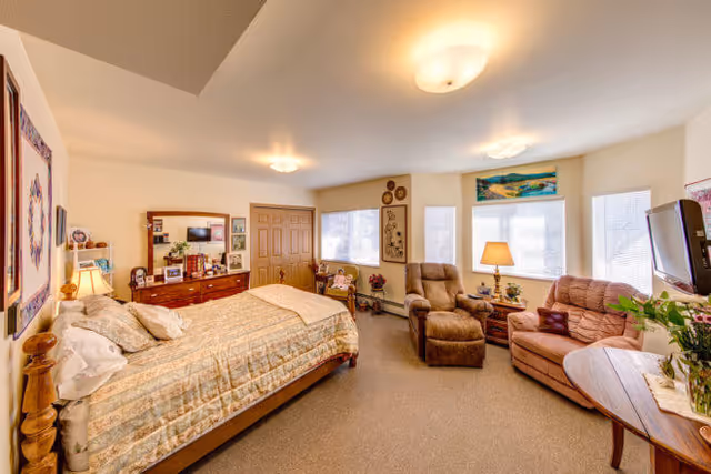 Furnished senior bedroom featuring a bed, dresser, two recliners, a TV and large windows.