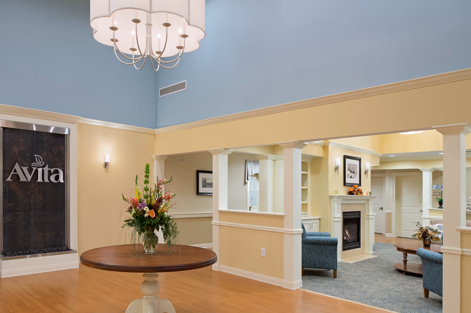 Bright senior living facility lobby with a round table holding a flower arrangement, seating area with a fireplace, and an 'Avita' sign on the wall.