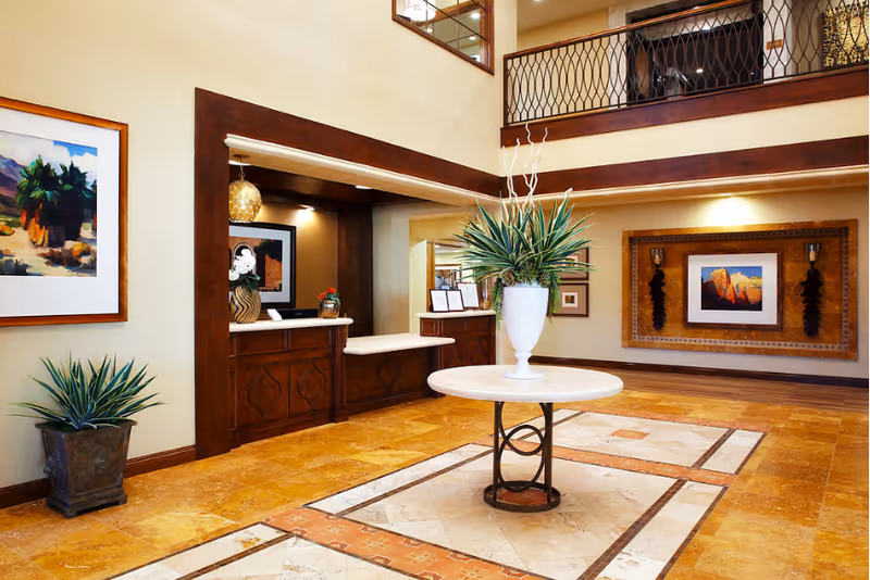 Spacious and well-lit interior lobby area with a round table in the center holding a large white vase with green plants. The floor has a decorative tile pattern, and the walls are adorned with framed artwork. There is a wooden reception desk area with a small flower arrangement and a two-story ceiling with a balcony railing above.