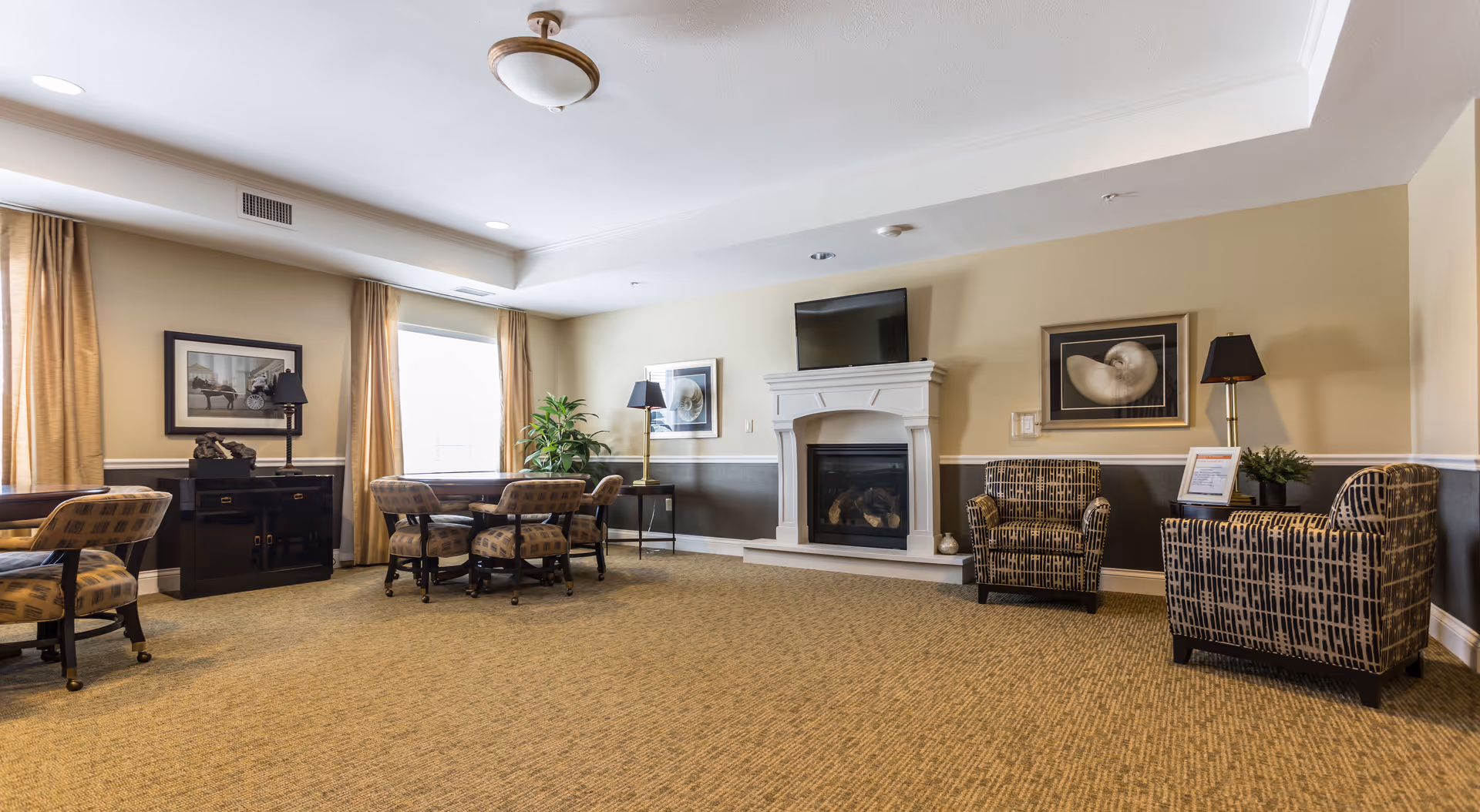 Spacious senior living communal lounge with seating areas, tables, a fireplace with TV above, and framed artwork.