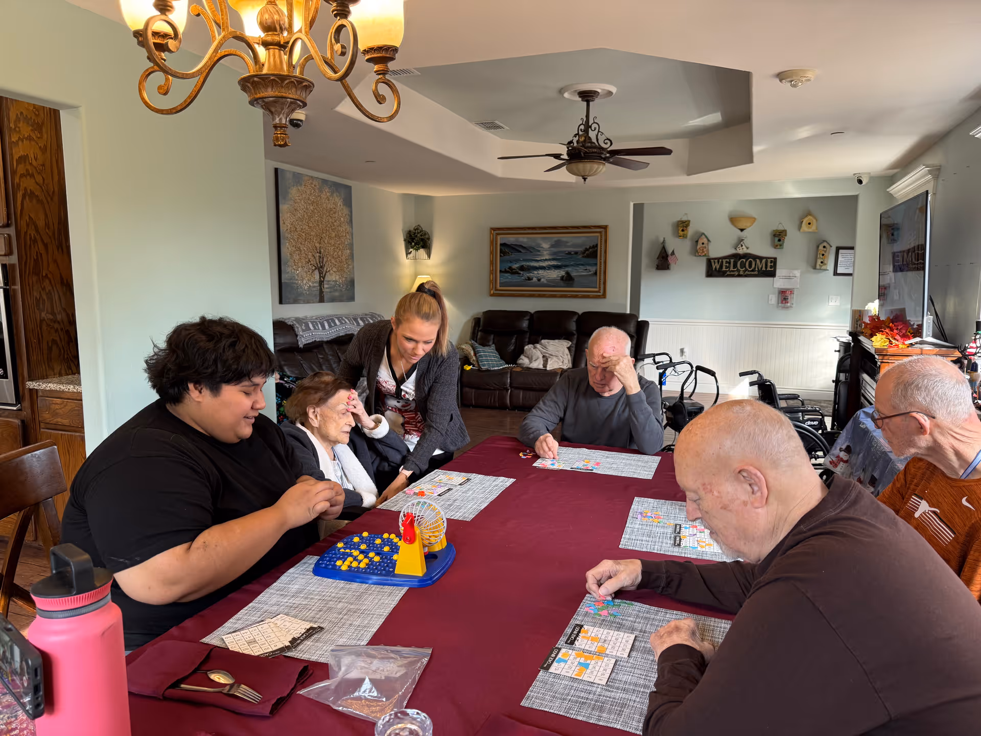 A group of elderly people and a caregiver sitting around a table playing a bingo game in a well-lit room with a chandelier and ceiling fan. The room has light blue walls, a brown leather couch, and decorative wall art including a 'WELCOME' sign and birdhouses.