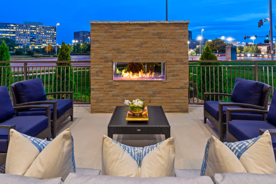 Outdoor patio area at dusk with a modern rectangular stone fireplace in the center. Surrounding the fireplace are cushioned wicker chairs with dark blue cushions and a black coffee table with a decorative centerpiece. In the background, there is a metal railing and a view of a cityscape with buildings and streetlights.