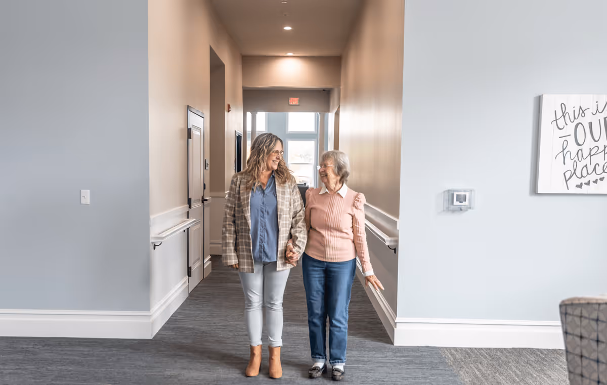 Two women, one younger and one elderly, walking hand in hand down a well-lit hallway in a senior living facility. The hallway has light-colored walls, handrails, and a carpeted floor. A decorative sign on the wall reads 'this is our happy place.'