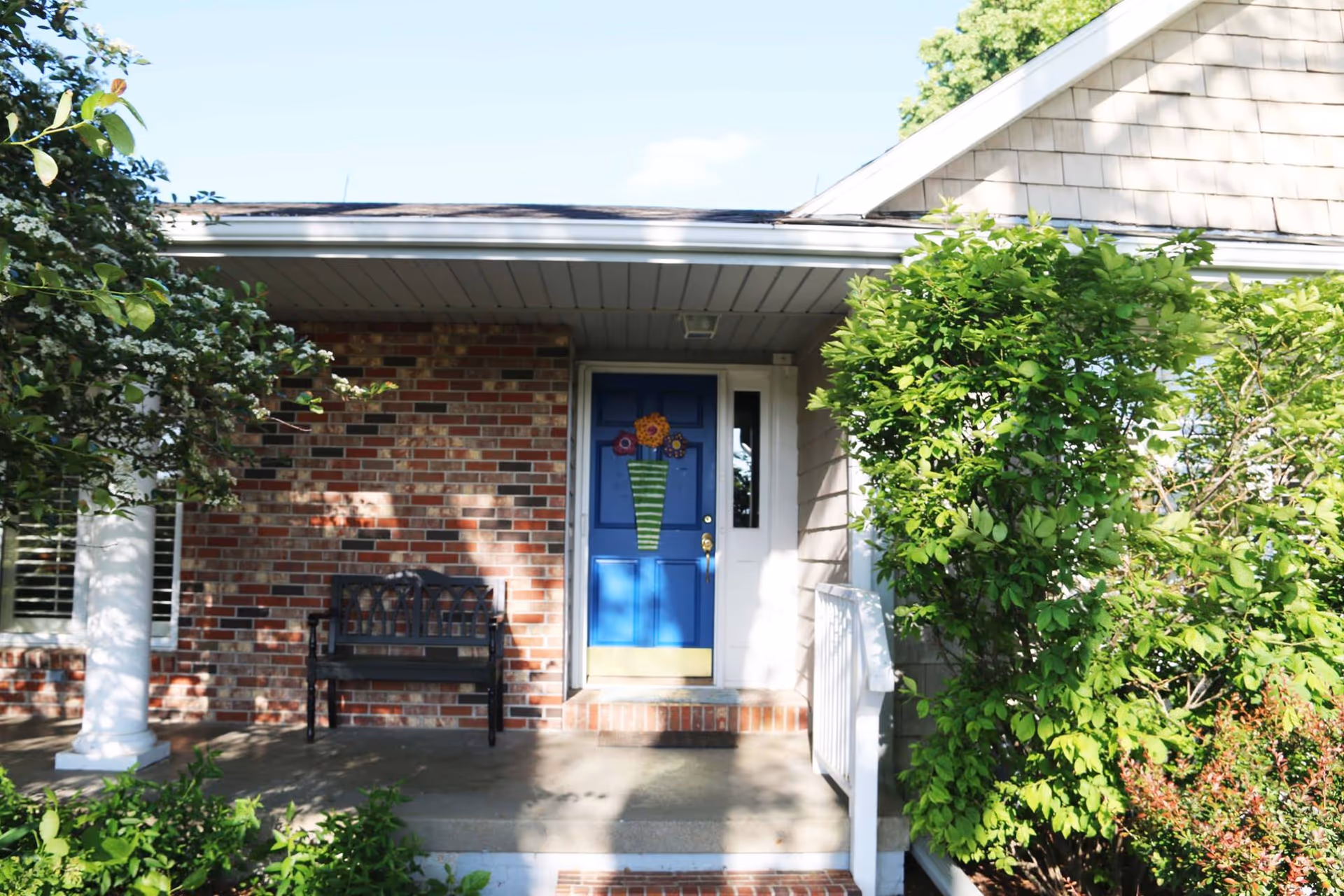 Front entrance of a house with a blue door decorated with a flower wreath, a black bench on the porch, brick walls, white columns, and green bushes surrounding the entrance.