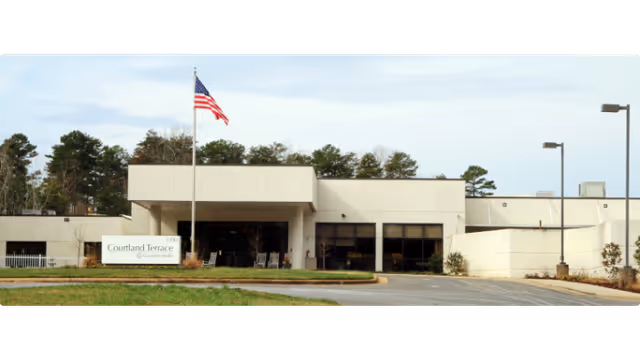Front exterior view of Courtland Terrace facility with an American flag on a flagpole in front, surrounded by trees and a driveway leading to the entrance.